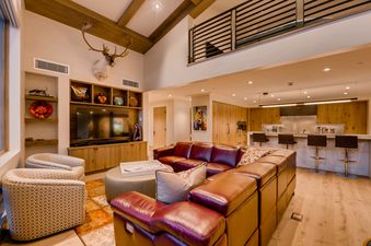 An expansive open-concept living area in The Gondola One Chalet, featuring a large leather sectional, two patterned accent chairs, a central ottoman, and a built-in media console with a TV, all set against a backdrop of a modern kitchen.