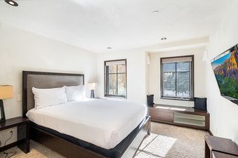 A bedroom with a white bed with a brown headboard, two windows and a flat-screen TV.