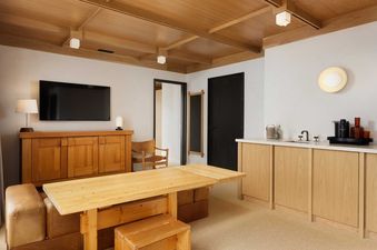 The image displays the comfortable and well-appointed living area of the Mollie Aspen Mollie Suite, featuring a large wooden dining table, a credenza with a TV, and a functional kitchenette with light wood cabinetry.