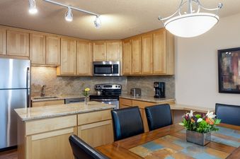 The kitchen at Vantage Point 102 features light wood cabinetry, stainless steel appliances including a refrigerator, microwave, and stove, and light granite countertops. The dining area has a dark wood table with a tiled inlay, black leather chairs, and a