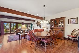 A rustic dining room with a round wooden table and Windsor-style chairs with plaid cushions, leading to a living area with a lake view.