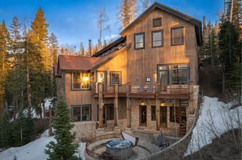 The Rocky Road Hideaway features stunning architecture with rustic wood and stone, surrounded by snow-covered terrain and tall pine trees under a clear blue sky.