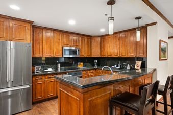 A well-lit, fully equipped kitchen with wooden cabinets, dark granite countertops, stainless steel appliances, and a central island with bar stools.