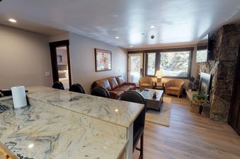 Manor Vail 163 is a spacious living room with a stone fireplace, a comfy leather couch, and two accent chairs. The room is decorated with a rustic theme and has a large window that overlooks the snowy mountains.