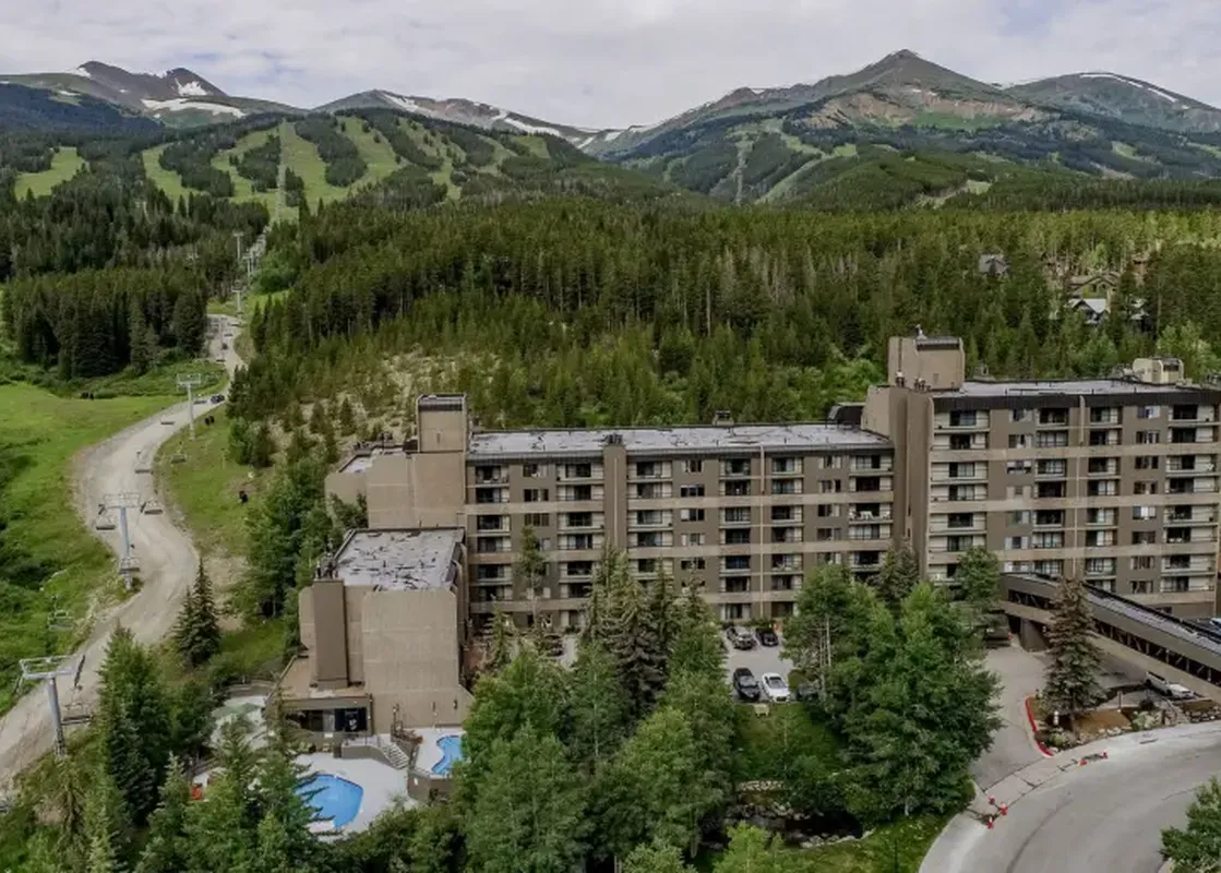 An aerial view of the Summit Suite Beaver Run Resort, showing its multi-story building, lush green surroundings, a winding road, and mountains with ski lifts in the background.