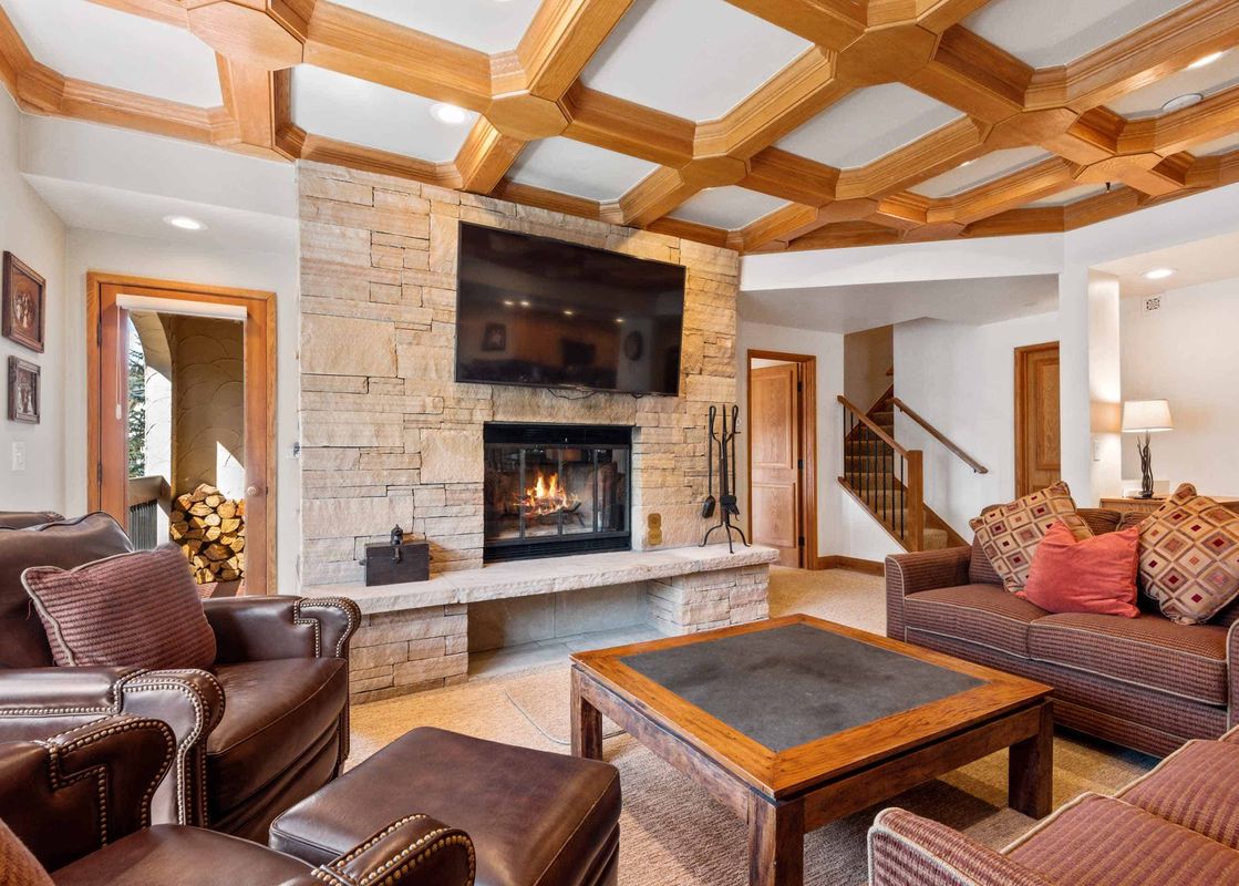 The living room of The Charter F3205 is designed with a coffered wooden ceiling and a stone fireplace, offering a cozy and inviting atmosphere.