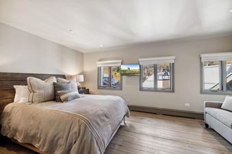 The primary bedroom features a king-sized bed, light-toned hardwood flooring, windows with blinds, a wall-mounted TV, and a bedside table with a lamp.