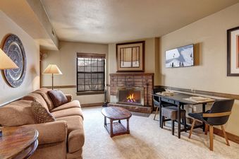A cozy living room in Copperbottom 207 with a warm fireplace, a brown sofa, a dining table, and a wall-mounted TV displaying a winter scene.