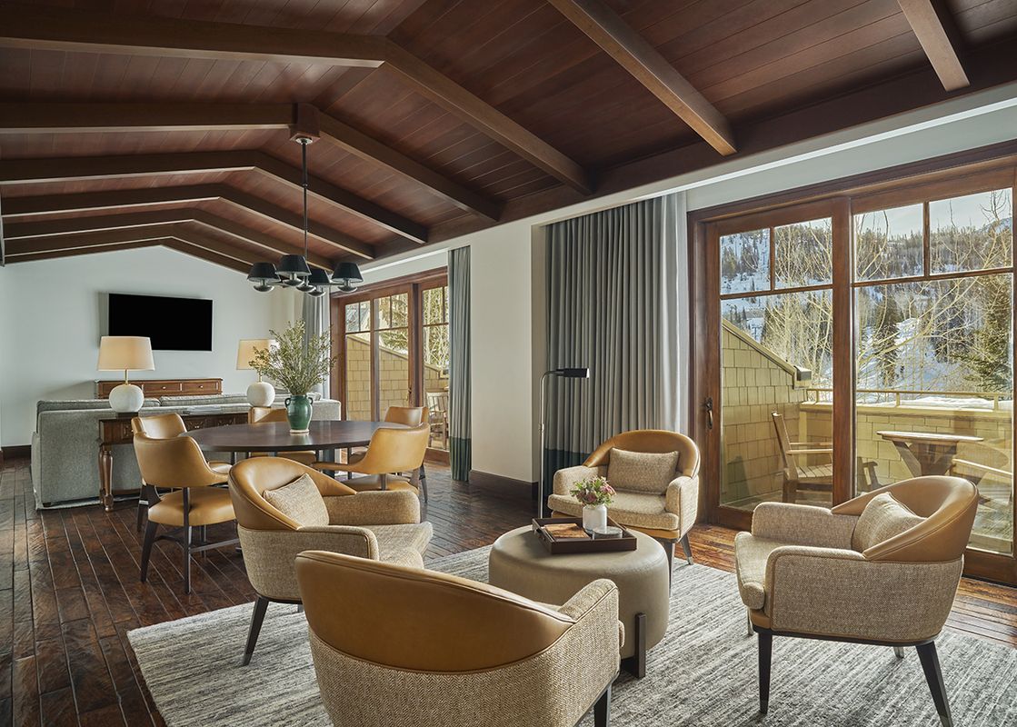 A luxurious and spacious living area in the Montage Two Bedroom Empire Suite features high vaulted wooden ceilings, an elegant dining area, a comfortable lounge area with an ottoman, and large glass doors opening to a snowy mountain view.