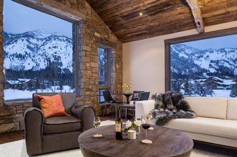 A luxurious living area in Four Pines 06 featuring cozy seating, a coffee table with wine, and large windows with stunning views of snow-covered mountains and pine trees.