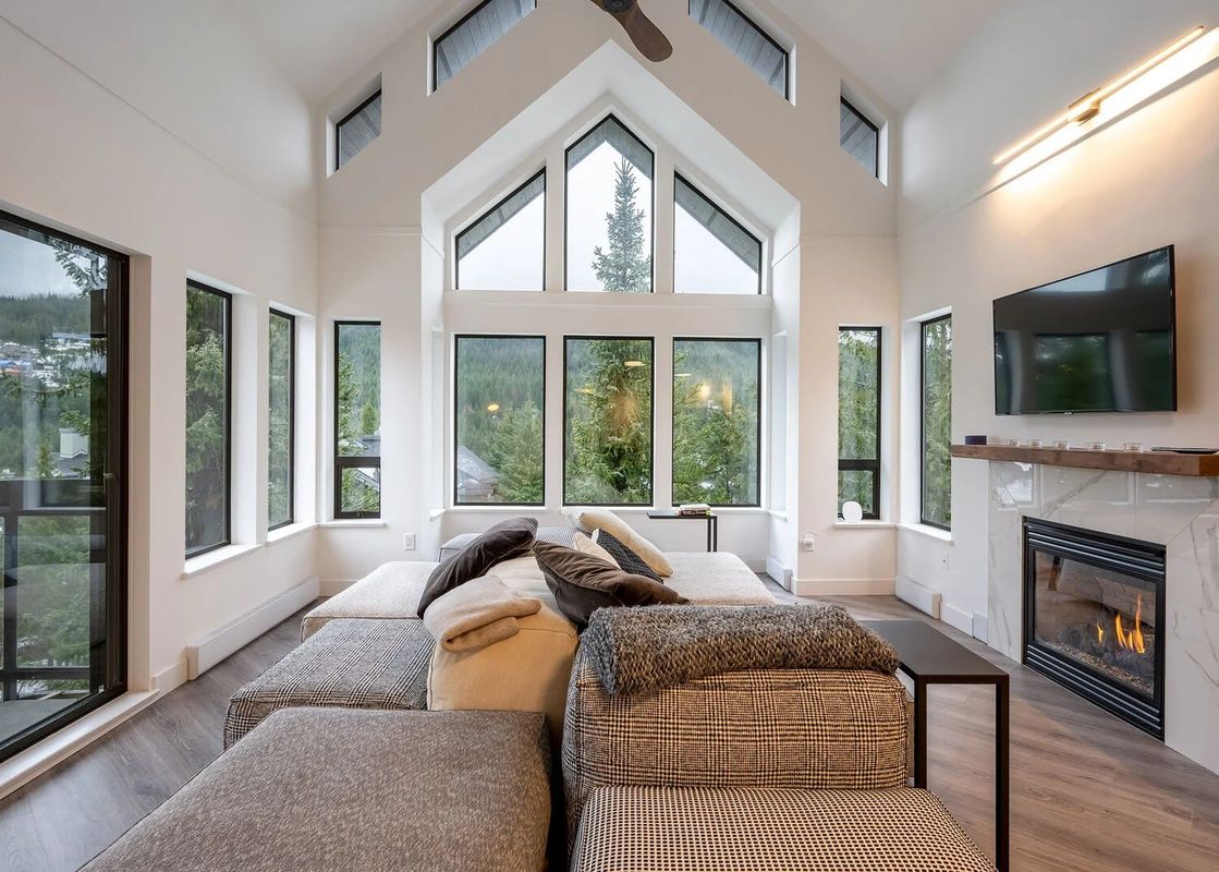 A modern and bright living area with high ceilings, a large sectional sofa, a fireplace, and large windows with forest views.