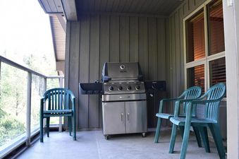 The image features a balcony with a stainless steel BBQ grill and three teal plastic chairs, set against a backdrop of trees.