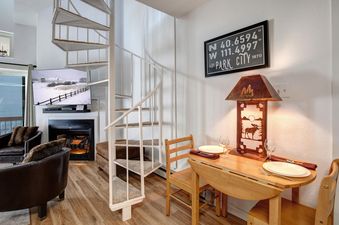 A dining nook with a wooden table and chairs is next to a spiral staircase, with a living area and fireplace in the background.