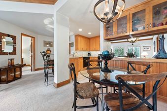 A dining table with a glass top and wooden chairs is set next to a kitchen with light wood cabinets and a pass-through bar.