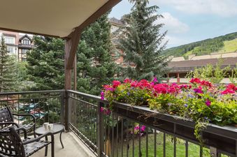 A private balcony at Montaneros 216, adorned with vibrant red and purple flowers in a planter box, features two chairs and a small table, offering a serene view of the village and distant mountains.