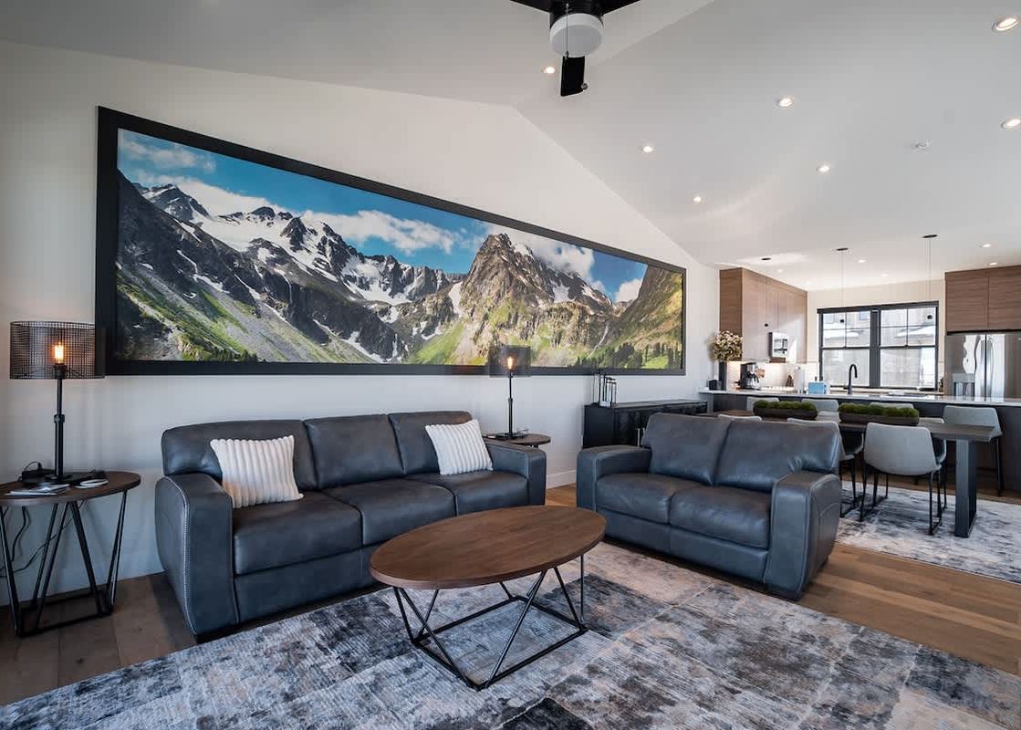 A spacious living room at Penthouse in the Pines, featuring comfortable sofas, a large mountain landscape artwork, and an open-concept kitchen with modern appliances and bar seating.