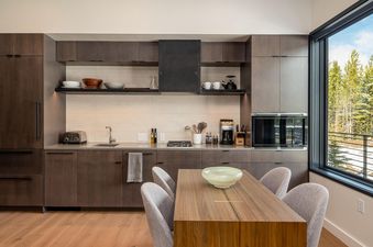 A modern kitchen and dining area with dark wood cabinetry, integrated appliances, and a dining table with chairs, featuring a large window with a view of a snowy forest.