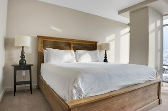 A serene king bedroom in Lowell 301A features a wooden bed, white linens, and elegant nightstand lamps, with a glimpse of a snowy landscape through the window.