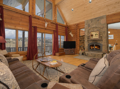 The living area at 2665 Golden Eagle Drive is furnished with comfortable sofas and features a large stone fireplace, large windows, and mounted animal skulls.