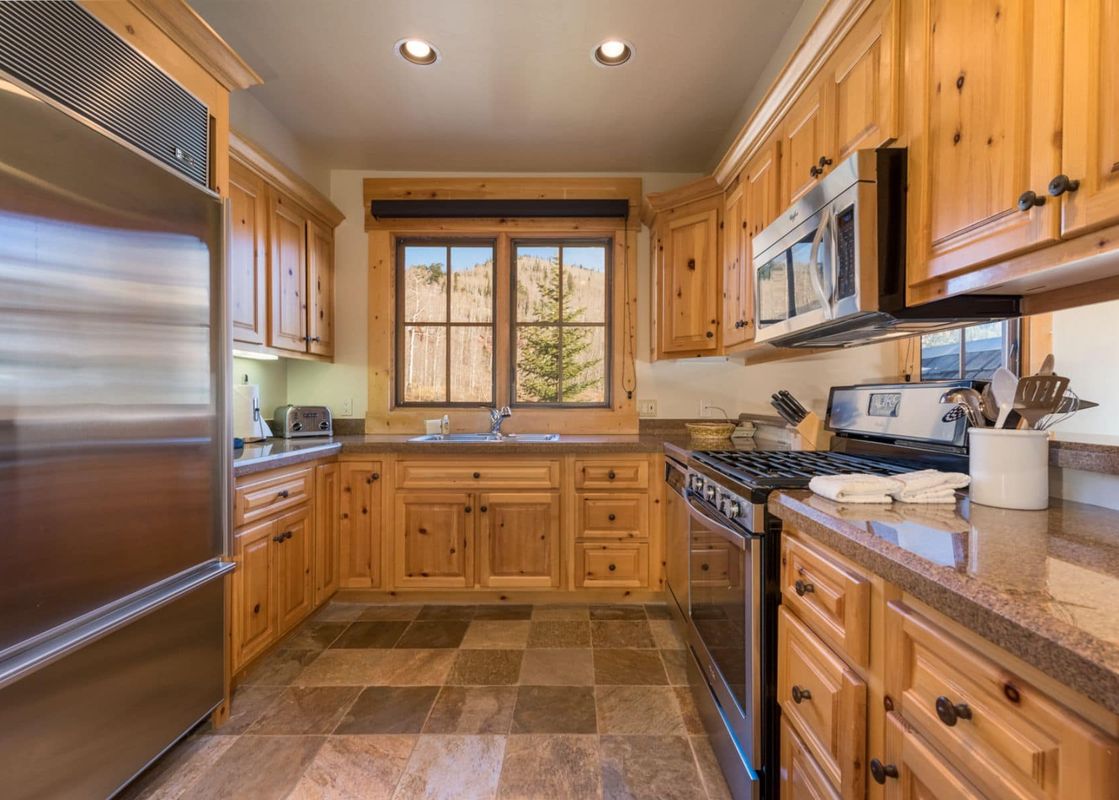 A rustic-chic kitchen with light wood cabinets, stainless steel appliances, stone countertops, and a window overlooking a mountainous, tree-filled landscape.