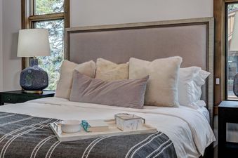 Bedroom at Moonlight Basin 6 Diversion Lane features a plush king-size bed with white linens and a grey throw blanket. The headboard is upholstered in a light grey fabric and accented with a decorative trim. There is a tray on the bed with a bowl and a bo