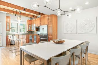 A modern dining room and kitchen with a long white table, grey chairs, wooden cabinetry, and unique lighting in Mountain Elegance 2835.