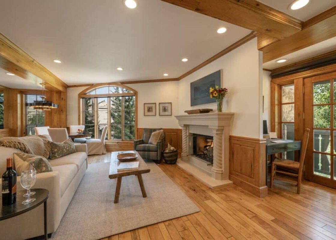 A luxurious living room at Chateau Beaver Creek Residence 203 with a stone fireplace, comfortable sofa, rustic coffee table, and large arch-top windows offering views of evergreen trees.