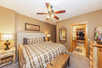 A spacious bedroom featuring a king-sized bed with plaid bedding, a wooden bench, a large mirror, and access to a games room.