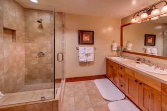 A bathroom with dual sinks, a shower with a glass door, a large mirror with lights above, and brown cabinets.