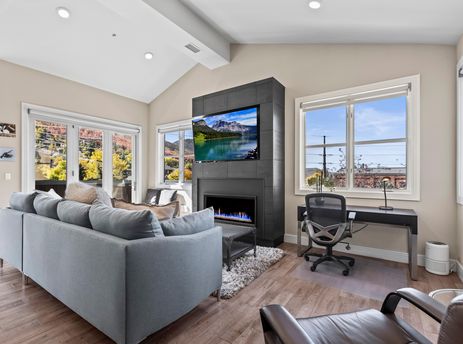 A modern and inviting living room featuring a grey sectional sofa, a fireplace with a mounted television, a dedicated desk area, and large windows revealing colorful autumn trees outside.