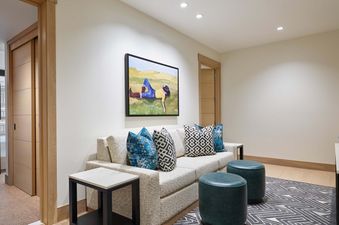 A den in the Cirque Two Bedroom Den Residence featuring a sofa with decorative pillows, two teal ottomans, a patterned rug, and a framed painting on the wall.