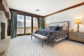 A master bedroom at Silver Strike Lodge 605 is elegantly designed with a blend of comfort and mountain-inspired aesthetics, featuring a spacious bed, a large fireplace, and a walk-out balcony that offers stunning views of snow-covered mountains.