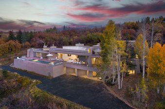 An aerial view captures the Eppley Estate, a modern luxury home with a swimming pool and outdoor terrace, surrounded by a vibrant autumn forest under a dramatic sky.