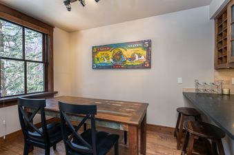 A dining area with a rustic wooden table, black chairs, and a large window offering a view of pine trees.