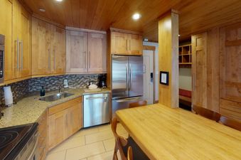 A kitchen with stainless steel appliances and wooden cabinets, a dining table with chairs and a view into another room with wooden cabinets and a bench.