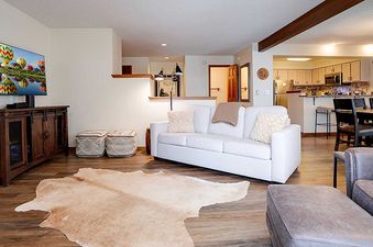 A wide view of the open-concept living room, kitchen, and dining area at Ranch at Steamboat RA203, featuring a white sofa, a large TV, a hide rug, and a modern kitchen.