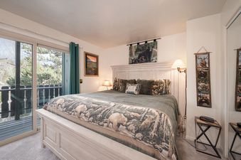 A cozy bedroom with a large bed, featuring a patterned comforter and pillows with nature-themed designs, has a sliding glass door leading to a balcony with a wooded view.