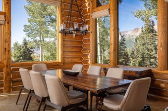 The dining area in Bear Lodge features a large wooden table and comfortable chairs, set against a backdrop of panoramic windows showcasing a lush mountain landscape.