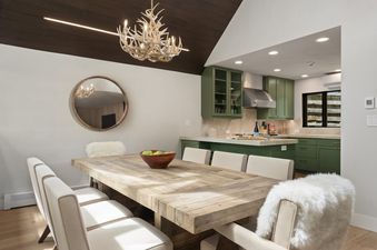 A dining room at Onyx Lodge with a rustic wooden table and upholstered chairs, leading into a kitchen with olive-green cabinets and modern appliances.