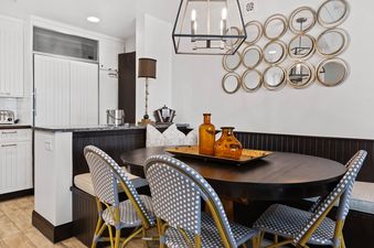 The dining nook features a dark wood oval table, stylish woven chairs, a modern chandelier, and a decorative wall art arrangement of gold-framed mirrors.