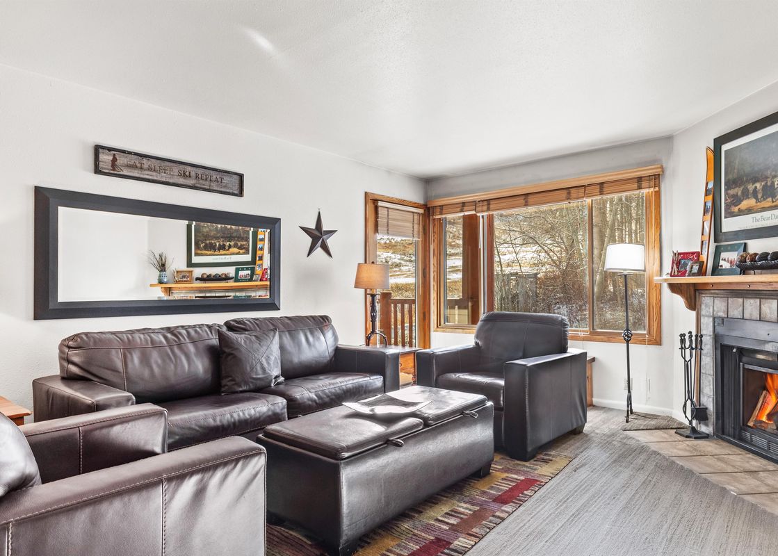 A cozy living room featuring dark leather furniture, a fireplace with a lit fire, and a large window showcasing a snowy outdoor scene.