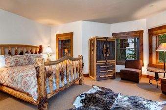 A rustic bedroom in The Moosehead Cabin features a log bed with floral bedding, a unique wooden armoire, and natural light from exterior view windows.