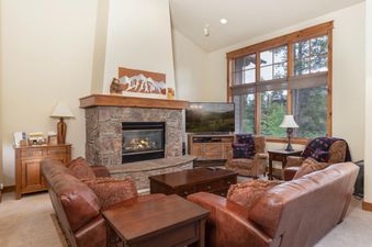 A cozy living room with a stone fireplace, comfortable brown leather sofas, and a large window looking out onto a lush green landscape.