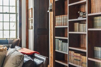 A rustic library at Five Trees Estate featuring a dark wooden built-in bookcase filled with vintage books, a comfortable leather sofa with plush pillows, and a large window providing natural light.
