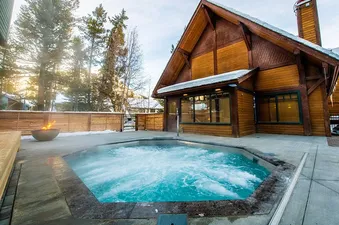 An outdoor hot tub with bubbling blue water is set in front of a rustic wooden lodge, surrounded by pine trees and a fire pit, hinting at a cozy, relaxing retreat.