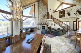 A spacious living room with a large stone fireplace, a dining table in the foreground, and a view of the mountains through large windows.