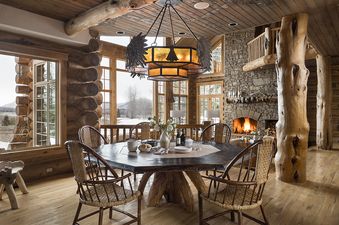 A rustic dining room at Mountain View Clear Creek features a large octagonal wooden table, wicker chairs, a unique chandelier, and a stone fireplace with a roaring fire, all set against a backdrop of snowy mountain views through expansive windows.