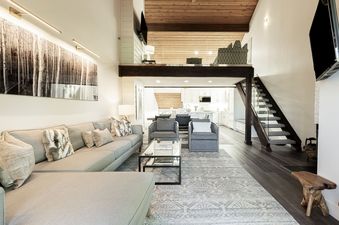 A modern living room with a large L-shaped sofa, two armchairs, a glass coffee table, and a nature-inspired artwork, illuminated by natural light and overhead fixtures.