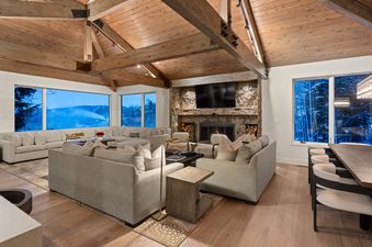 A wide shot of Casa de Gondola's expansive living room with vaulted wooden ceilings, a stone fireplace, and large windows showcasing a snowy mountain view.