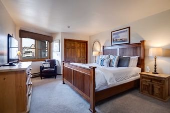 A spacious bedroom at Chateau Chamonix CX222 featuring a king-sized bed, wooden dresser with a TV, a cozy armchair, and a window with an exterior view.